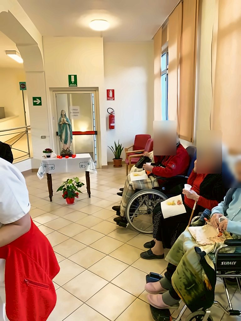 Ospiti seduti in un corridoio davanti a una statua della Madonna con fiori e lumini; i volti sono oscurati