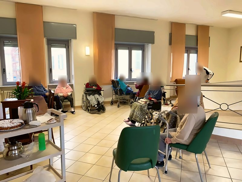 Ospiti seduti nel salone luminoso nel pomeriggio, con un carrello della merenda in primo piano; i volti sono oscurati